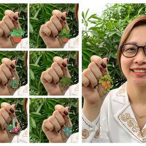 Op de afbeelding: Een set van zes emaillepins met verschillende hangende planten in geometrische terrariums. De pins zijn goudkleurig met groene, bruine en roze details.