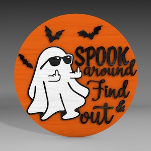 Op de afbeelding: Een rond, oranje houten bord met een zwarte geest die een zonnebril draagt en zijn middelvinger opsteekt. Het bord zegt "Spook around find out &" in zwarte letters.