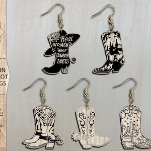 May include: Four pairs of laser cut wooden earrings shaped like cowboy boots. The boots have different designs, including a boot with a cactus, a boot with a mountain scene, and a boot with the text "Real Women Wear Cowboy Boots".
