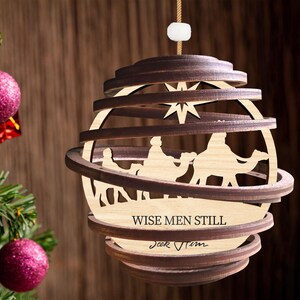 Puede incluir: Un adorno navide&ntilde;o de madera con forma de esfera con un dise&ntilde;o en capas. El adorno presenta una escena de los tres reyes magos montando camellos, con una estrella sobre ellos. El texto "Wise Men Still Seek Him" est&aacute; inscrito en el adorno.