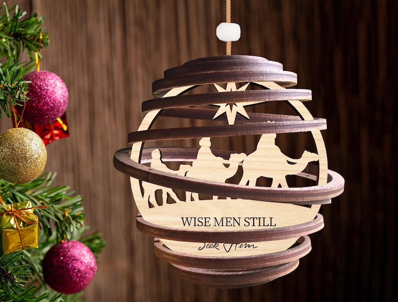 Puede incluir: Un adorno navide&ntilde;o de madera con forma de esfera con un dise&ntilde;o en capas. El adorno presenta una escena de los tres reyes magos montando camellos, con una estrella sobre ellos. El texto "Wise Men Still Seek Him" est&aacute; inscrito en el adorno.