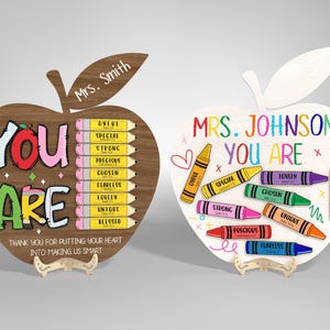 May include: Two wooden apple-shaped plaques for teachers. One is brown with colorful text and pencils, the other is white with crayons. Both say "You Are" and have inspirational words. One says "Mrs. Smith", the other "Mrs. Johnson".