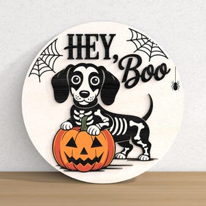 以下が含まれることがあります： ハロウィーンの丸い木製サイン。スケルトンのコスチュームを着たダックスフンド犬が、オレンジ色のカボチャを持っています。サインには「HEY, Boo」と書かれており、クモの巣とクモが描かれています。犬は黒い耳と白い顔をしています。