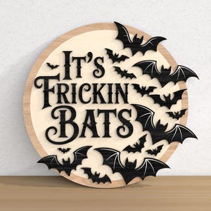 以下が含まれることがあります： 「It's Frickin' Bats」の文字が黒で書かれた丸い木製の看板。黒いコウモリのシルエットが、濃い木材の縁取りのある明るい木材の背景にテキストを囲んでいます。ハロウィーンの装飾に最適です。