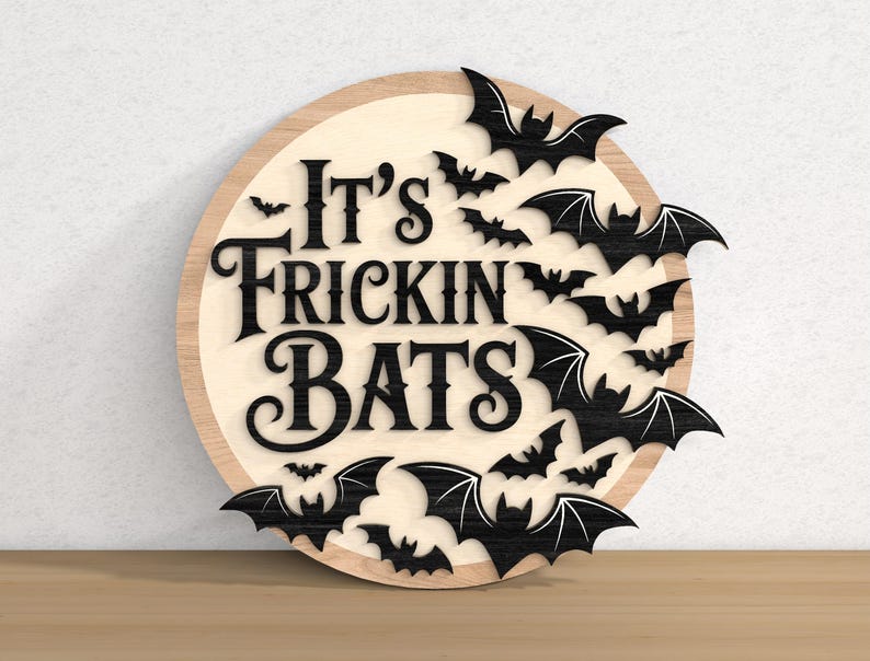 以下が含まれることがあります： 「It's Frickin' Bats」の文字が黒で書かれた丸い木製の看板。黒いコウモリのシルエットが、濃い木材の縁取りのある明るい木材の背景にテキストを囲んでいます。ハロウィーンの装飾に最適です。