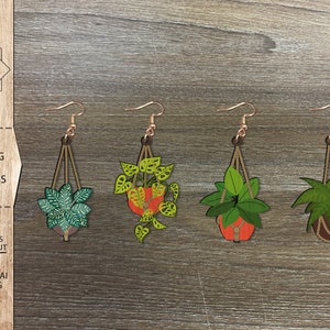 Op de afbeelding: Vier paar lasergesneden houten oorbellen met hangende planten in potten. De planten zijn groen en hebben verschillende bladvormen. De potten zijn bruin, oranje en rood. De oorbellen zijn een leuke en trendy accessoire.
