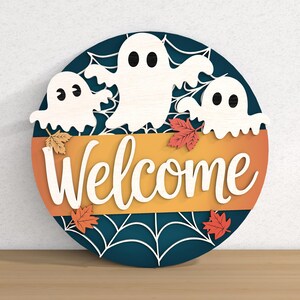 以下が含まれることがあります： 3つの白い幽霊、オレンジと黄色のアクセント、筆記体で「Welcome」の文字が入った、ハロウィーンをテーマにした丸い木製サイン。秋の葉とクモの巣のデザインも特徴です。
