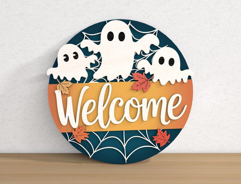 以下が含まれることがあります： 3つの白い幽霊、オレンジと黄色のアクセント、筆記体で「Welcome」の文字が入った、ハロウィーンをテーマにした丸い木製サイン。秋の葉とクモの巣のデザインも特徴です。