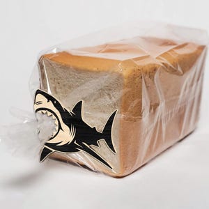 May include: A loaf of white bread in a clear plastic bag, secured with a wooden shark-shaped clip. The shark clip is black and natural wood, with a detailed open mouth design.