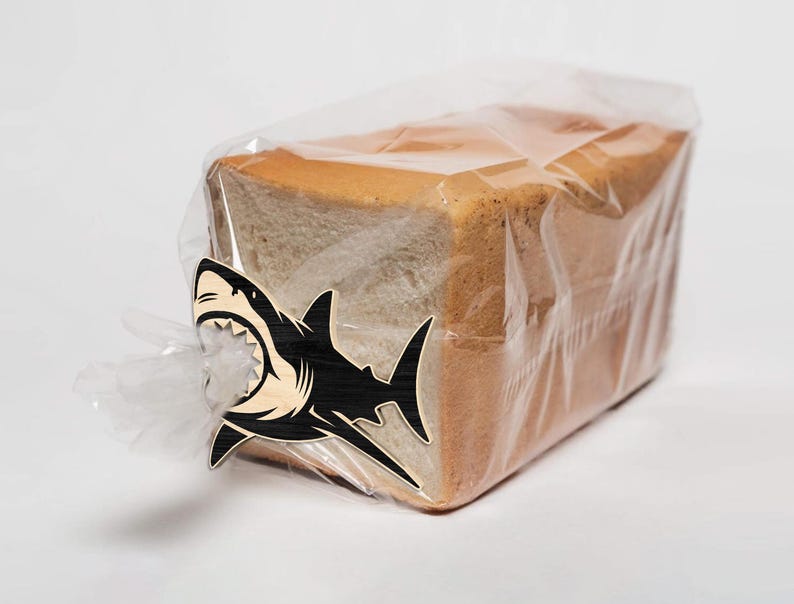 May include: A loaf of white bread in a clear plastic bag, secured with a wooden shark-shaped clip. The shark clip is black and natural wood, with a detailed open mouth design.