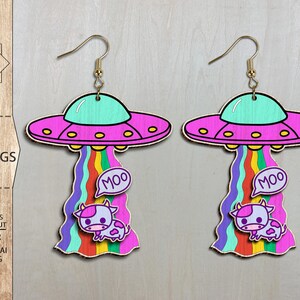 Könnte beinhalten: Ein Paar rosa und lila UFO-Ohrringe mit einem Regenbogen-Schweif und einer Comic-Kuh mit dem Wort "MOO" in einer Sprechblase. Die Ohrringe sind zum Laserschneiden geeignet.