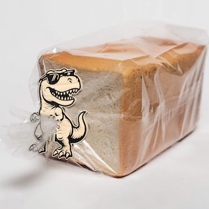 May include: A loaf of sliced white bread in a clear plastic bag. A wooden dinosaur wearing sunglasses is attached to the bag. The bread has a golden-brown crust and a soft, white interior. The dinosaur is light brown with black details.