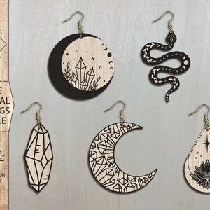May include: Set of five laser cut wooden earrings featuring mystical designs. The earrings include a crescent moon with crystals, a snake with a crescent moon, a crystal, a crescent moon with crystals, and a teardrop with a lotus flower and a star.