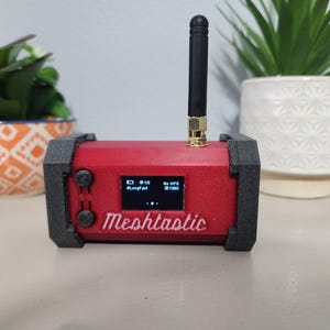 May include: A red, rectangular device with a black antenna and a small screen displaying text and numbers. The device is labeled "Meshtactic".