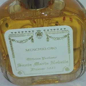 Puede incluir: Un frasco de perfume de vidrio transparente con tapón y etiqueta dorados. La etiqueta dice "Muschio Oro" y "Officina Profumo Santa Maria Novella Firenze 1221". El perfume en su interior es de color ámbar.