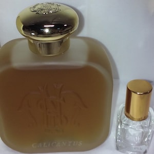 Op de afbeelding: Een parfumfles en een kleine monsterfles. De grote fles is vierkant met een gouden dop en het woord "CALICANTUS" op de voorkant. De kleine fles heeft een gouden dop.