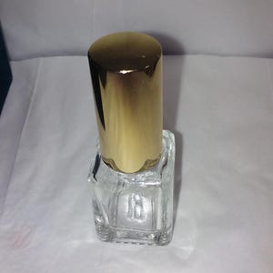 Może przedstawiać: Przezroczysta, kwadratowa szklana butelka perfum z pozłacaną nakrętką. Butelka jest pusta i stoi pionowo na białej powierzchni. Nakrętka jest cylindryczna i błyszcząca.