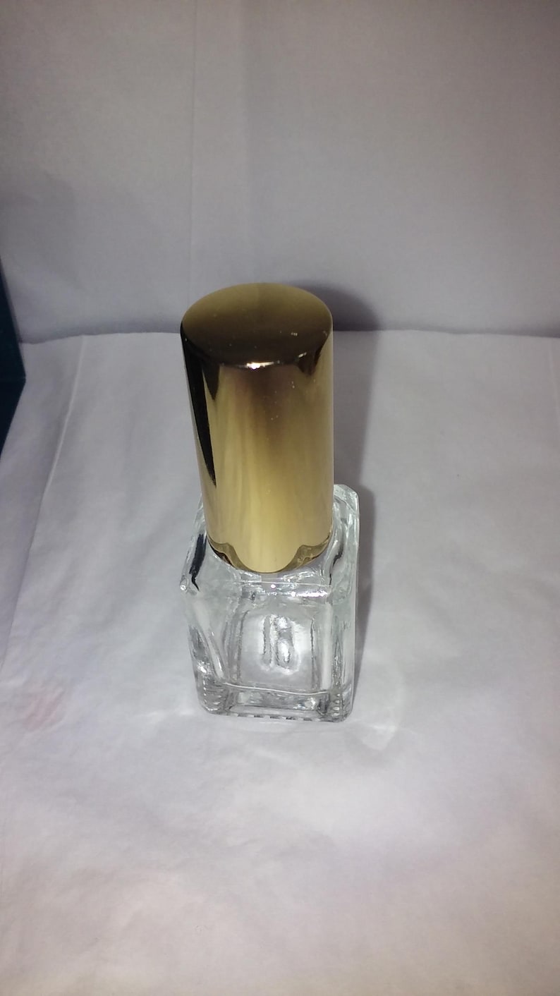 Może przedstawiać: Przezroczysta, kwadratowa szklana butelka perfum z pozłacaną nakrętką. Butelka jest pusta i stoi pionowo na białej powierzchni. Nakrętka jest cylindryczna i błyszcząca.