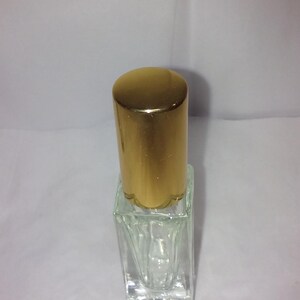 Może przedstawiać: Przezroczysta, pusta butelka perfum ze złotą nakrętką. Butelka ma kwadratową podstawę i cylindryczną nakrętkę. Nakrętka ma karbowaną krawędź. Butelka stoi na białej powierzchni.