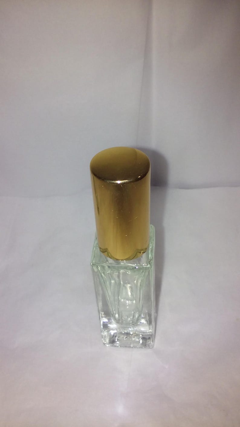 Może przedstawiać: Przezroczysta, pusta butelka perfum ze złotą nakrętką. Butelka ma kwadratową podstawę i cylindryczną nakrętkę. Nakrętka ma karbowaną krawędź. Butelka stoi na białej powierzchni.