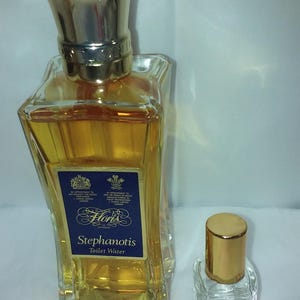 May include: Two glass perfume bottles. The larger bottle has a gold cap and a blue label that reads "Stephanotis Toilet Water". The smaller bottle is clear with a gold cap. Both bottles are filled with a golden liquid.
