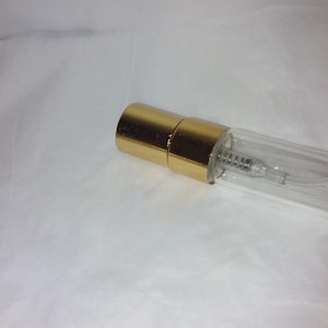 Może przedstawiać: Przezroczysty szklany flakon perfum ze złotą nasadką. Butelka ma mały, cylindryczny kształt z widoczną metalową sprężyną w środku. Nasadka ma błyszczący, metaliczny złoty kolor.