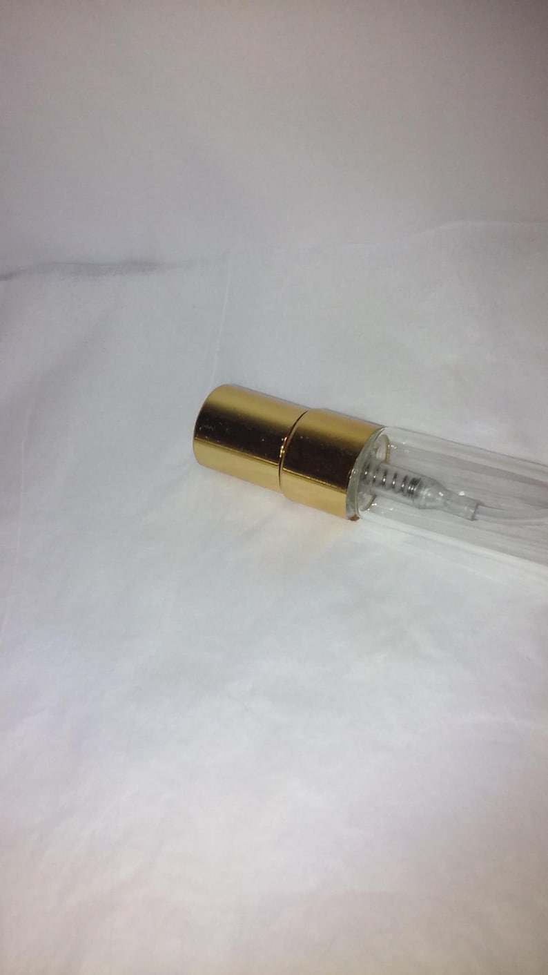 Może przedstawiać: Przezroczysty szklany flakon perfum ze złotą nasadką. Butelka ma mały, cylindryczny kształt z widoczną metalową sprężyną w środku. Nasadka ma błyszczący, metaliczny złoty kolor.