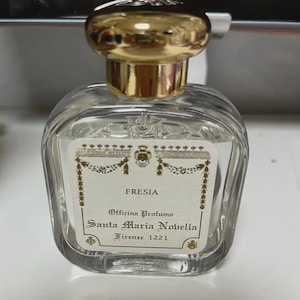 May include: A clear, square perfume bottle with a gold cap. The label reads "FRESIA" and "Officina Profumo Santa Maria Novella Firenze 1221". The bottle has a classic, elegant design.