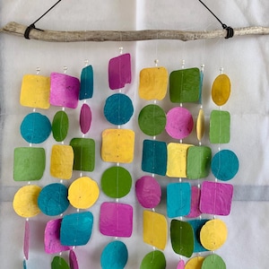 May include: A colorful wall hanging featuring various shapes in shades of yellow, pink, green, and blue. The shapes are suspended from a natural wooden branch with a black cord. The shapes include squares, circles, and oblongs.