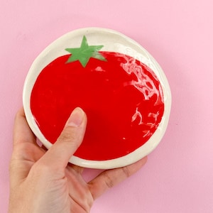 Puede incluir: Un plato de cerámica blanca con forma de tomate con una hoja verde en la parte superior. El plato es rojo y brillante.