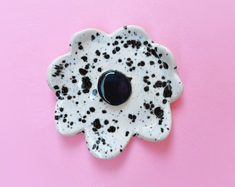May include: A white ceramic dish with black speckles and a black center. The dish is shaped like a flower with rounded edges.
