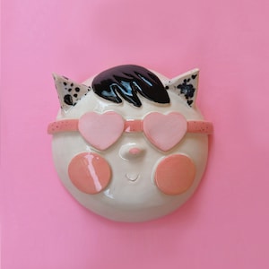 Könnte beinhalten: Keramik-Wandbehang in Form eines Katzen-Gesichts. Die Katze hat schwarzes Haar, rosa herzförmige Sonnenbrillen und rosa Wangen. Die Ohren haben schwarze Flecken. Der Hintergrund ist einfarbig rosa.