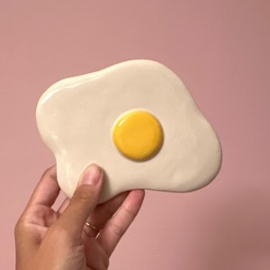 Può includere: Un piatto in ceramica a forma di uovo fritto, con una base color crema e un tuorlo giallo brillante. Il piatto è tenuto su uno sfondo rosa, mostrando il suo design giocoso. Ideale per l'arredamento della cucina.
