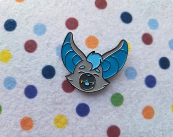 May include: A gray and blue enamel pin featuring a cartoon bat with open wings. The bat has a gray face, blue wings, and a small tuft of blue hair. The pin is set against a white background with colorful polka dots.