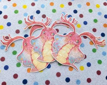 May include: Three pink and coral-colored creature stickers with detailed illustrations. The stickers have a layered design with a light yellow underbelly and a textured, iridescent wing-like section. The background is a white fabric with colorful polka dots.
