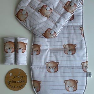 May include: A baby stroller accessory set with a white fabric, brown bear pattern, and pink stripes. The set includes a stroller liner, two strap covers, and a headrest. A wooden sign with the text "Buby by Em Louise" is also visible.