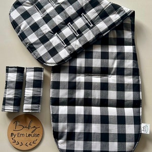 May include: Black and white checkered stroller liner with matching armrests. The liner is padded and has a rounded shape. The armrests are rectangular and have a black and white checkered pattern. The liner has a tag that says "Baby By Em Louise".