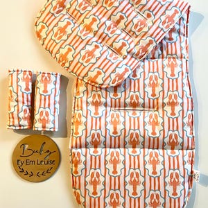 May include: A stroller liner and two matching harness strap covers featuring an orange lobster pattern on a white background with light blue vertical stripes. A round wooden plaque is included with the text Baby By Em Louise.