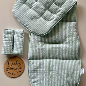 May include: A light green and white striped stroller liner with matching harness strap covers. A round wooden plaque is included with the text "Baby By Em Louise" and a decorative leaf graphic. The fabric features a textured seersucker pattern.