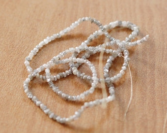 May include: A strand of gray diamond beads strung on a thin white thread. The beads are small and irregular in shape, creating a delicate and textured look.