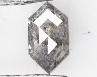 Może przedstawiać: Sześciokątny, szary diament trzymany przez metalową pęsetę. Diament ma geometryczny, biały wzór pośrodku. Kamień jest fasetowany i odbija światło, ukazując jego przejrzystość i szlif. Tło jest jednolicie białe.