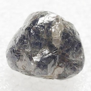 May include: A rough, uncut diamond with a dark gray and translucent appearance. The stone has an irregular, rounded shape with a textured surface. The diamond reflects light, showcasing its natural facets.