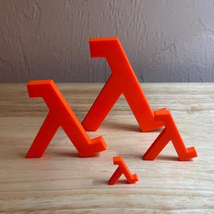 May include: Orange Lambda symbols in various sizes are arranged on a wooden surface. The symbols are 3D printed and have a solid, geometric design. The background is a textured, neutral-toned wall.