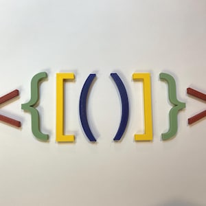 May include: Colorful wooden programming symbols arranged on a white surface. The symbols include angle brackets, curly braces, square brackets, and parentheses in shades of red, green, yellow, and blue.