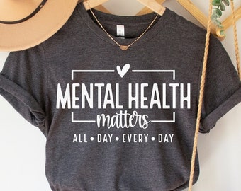 以下が含まれることがあります： 「MENTAL HEALTH matters ALL DAY EVERY DAY」の文字が入った濃いグレーのTシャツが、デニムショートパンツ、ベージュの帽子、マクラメバッグと一緒に表示されています。 Tシャツは木製のハンガーにかかっています。