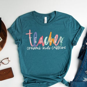 May include: Teal t-shirt with the word "teacher" in colorful letters, with "crayons, kids, caffeine" below. The shirt is styled with blue jeans and white sneakers. Other accessories include glasses, a small wallet, and a decorative wooden tray.