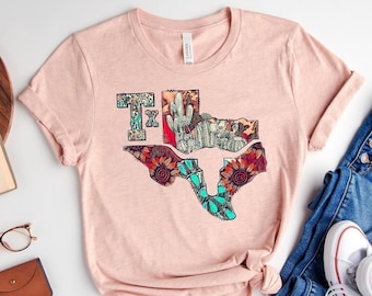 May include: A light pink t-shirt featuring a colorful Texas-shaped graphic with cacti, flowers, and the letters "TX". The shirt is paired with blue denim jeans, white sneakers, and accessories like a book, glasses, and a small brown wallet.