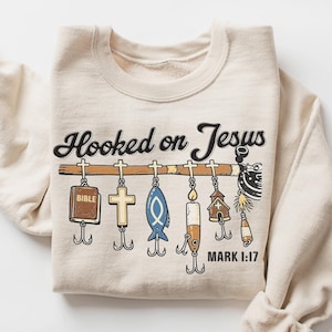 May include: Sand-colored sweatshirt with the text "Hooked on Jesus" above a fishing lure design. The lures include a Bible, cross, fish, and other religious symbols. The text "MARK 1:17" is printed below the lures.