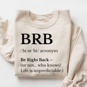 May include: A beige sweatshirt with the acronym "BRB" printed in black, along with its definition: "Be Right Back." The text also includes the phrase "Life is unpredictable." The sleeves are folded, and the sweatshirt is on a white surface.