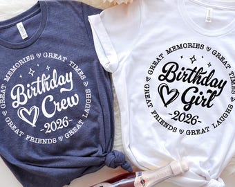 May include: Two t-shirts, one navy blue and one white, with the text "Birthday Crew" and "Birthday Girl" in a circular design. The design includes a heart, stars, and the year "2026". A bottle of sparkling wine is in the foreground.